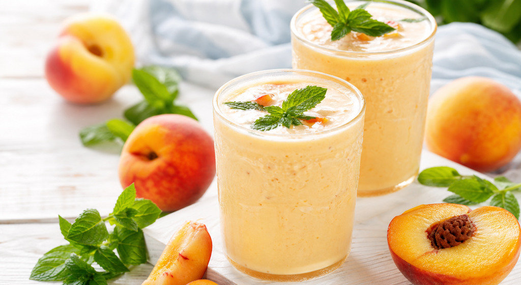 Zwei Gläser Pfirsich-Lassi mit Minze, frischen Pfirsichen und Minzblättern auf hellem Holztisch
